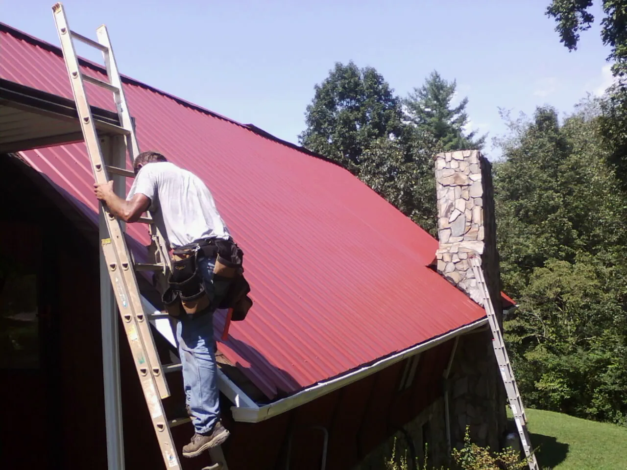 Roofer on a ladder beside a red metal roof and stone chimney — Soffit & Fascia Repair by Roofing Brothers in Mediapolis